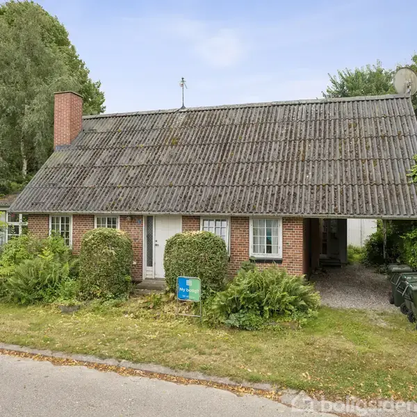 Et rødt murstenshus med gråt tegltag står omgivet af grønt buskads og træer. Huset har en skorsten og en antenne på taget. En affaldsspand står ved siden af indkørslen.