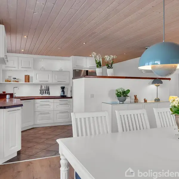 Et lyst køkken-udstyr. Hvide skabe og bordplader pryder rummet. En spisebordsgruppe med blomsterdekoration står midt i lokalet. Loftet er træbeklædt med indbyggede spotlys.