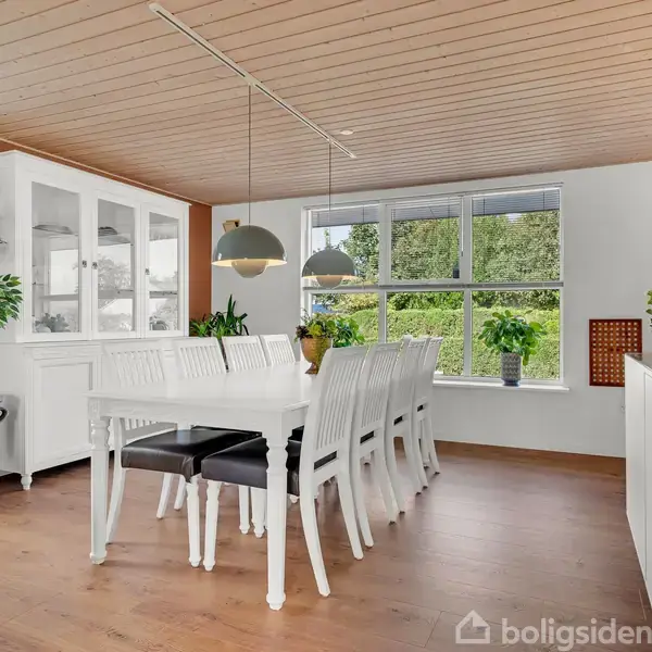 Hvidt spisebord med otte stole står i et lyst rum med store vinduer og planter. Hængende lamper over bordet skaber hyggelig belysning. Skab i hjørnet indeholder glasartikler.