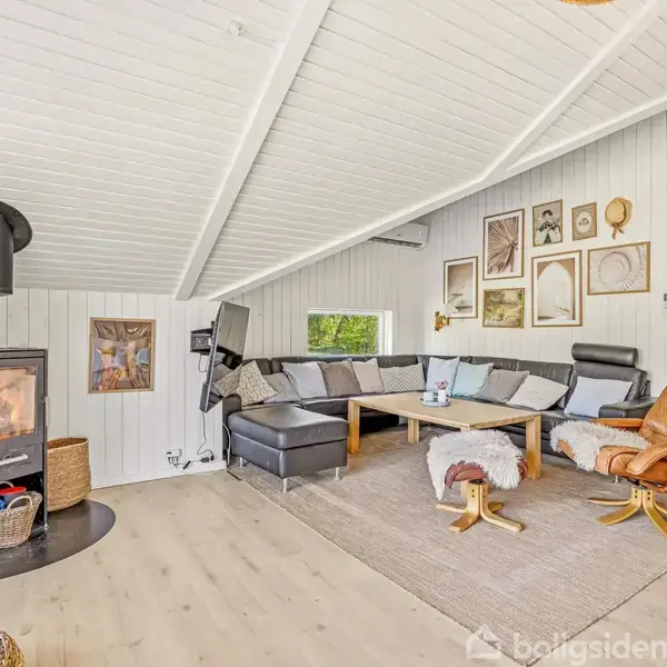 En stue med en brændeovn til venstre, hvor ilden brænder. En sort hjørnesofa står ved et lavt sofabord på et lys tæppe. Væggene har hylder og billeder.