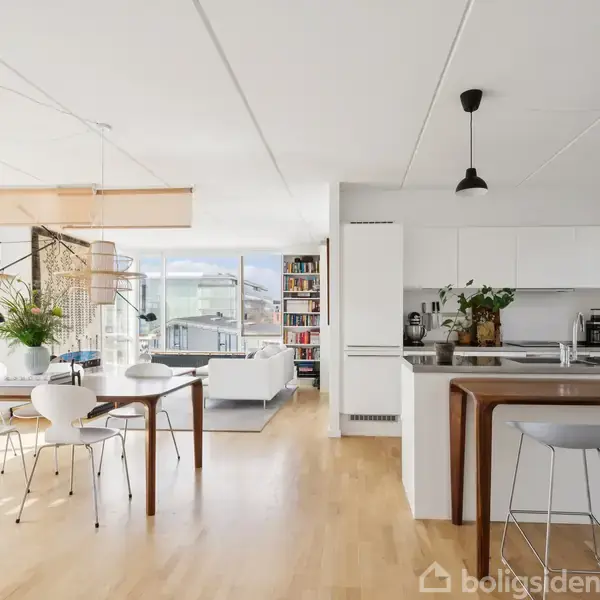 Et moderne køkken-alrum med hvide skabe og en køkkenø står centralt og omgives af spisebord med stole og en stue med sofaer. Store vinduer bringer naturligt lys ind.