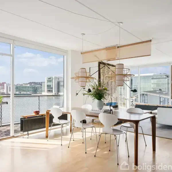Lys spiseplads med seks stole omkring bord i åben stue. Store vinduer med udsigt over byen. Hvid sofa i baggrunden nær bogreoler og balkon med udsigt.