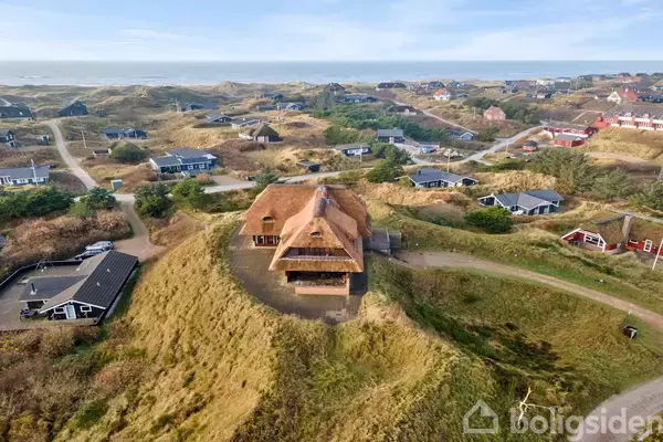 Et stort hus med stråtag står på en bakke omgivet af klitter. Flere mindre huse spredt i området. Havet ses i baggrunden.