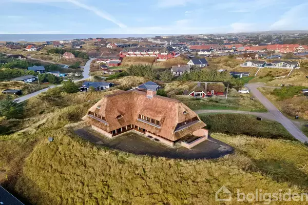 Hus med stråtag står på en bakke omgivet af græsarealer; i baggrunden ses en bydel med spredte huse og veje.
