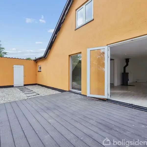 Et hus med orange facade og åbne terrassedøre fører til en træterrasse. Indendørs ses et rum med en brændeovn. Grusbelagt sti og græs er omkring huset.