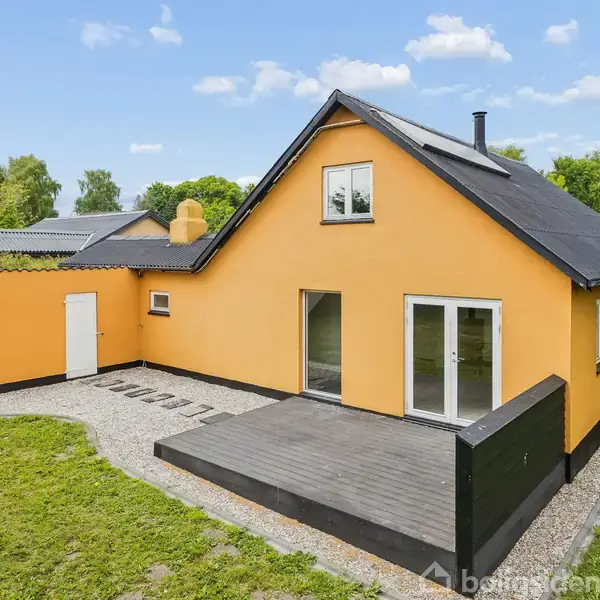Et gulmalet hus står roligt med en terrasse foran, omgivet af en grøn græsplæne og træer. Huset har en sort tagbelægning og flere vinduer.