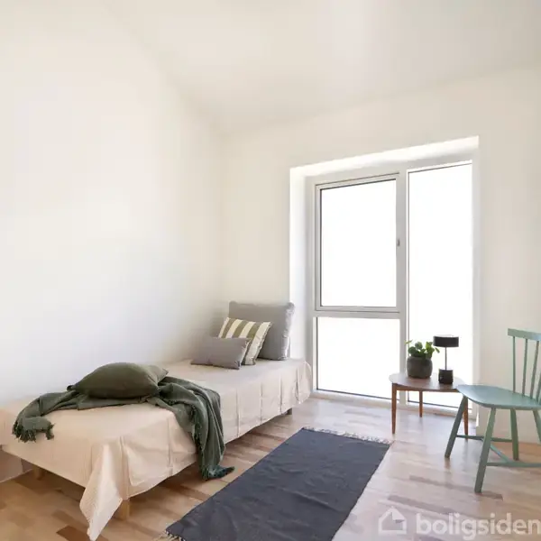 En enkeltseng med puder og tæpper står op mod væggen. Ved siden af er en grøn stol og et lille bord med en plante og lampe. Rummet er lyst og minimalistisk.