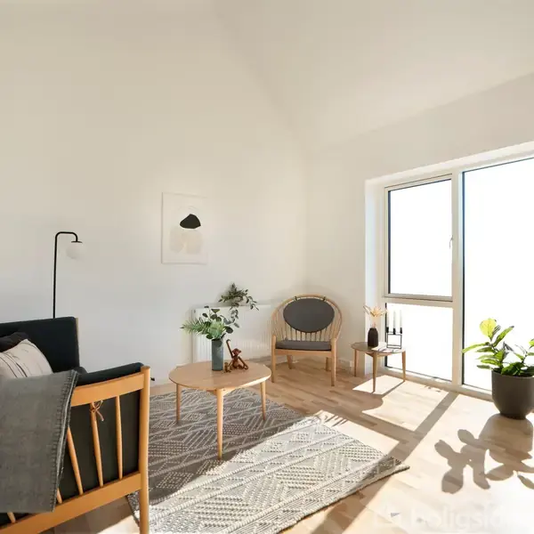Sofa med tæppe stående. Sofaen flankeres af et bord med blomster og en stol. Store vinduer lukker lys ind i rummet med træmøbler og en gulvplante.