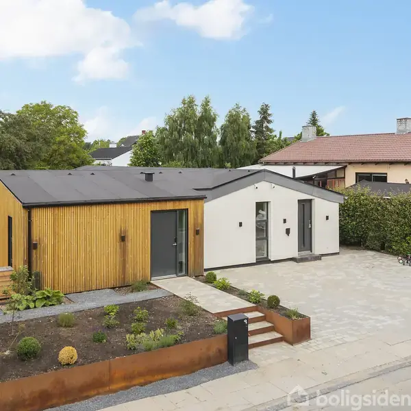 Et moderne hus med træbeklædning og hvide vægge står forrest på en stille villavej. Foran huset er en nydelig have og en brolagt indkørsel.