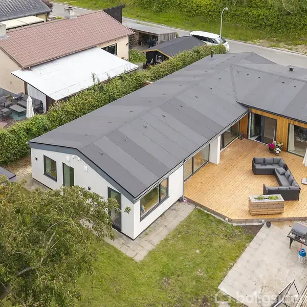 Modern enkeltfamiliehus med fladt tag ses fra fugleperspektiv. Huset har en stor terrasse med møbler og parasol. Omkringliggende grønne områder og en vej tæt ved bidrager til den rolige atmosfære.