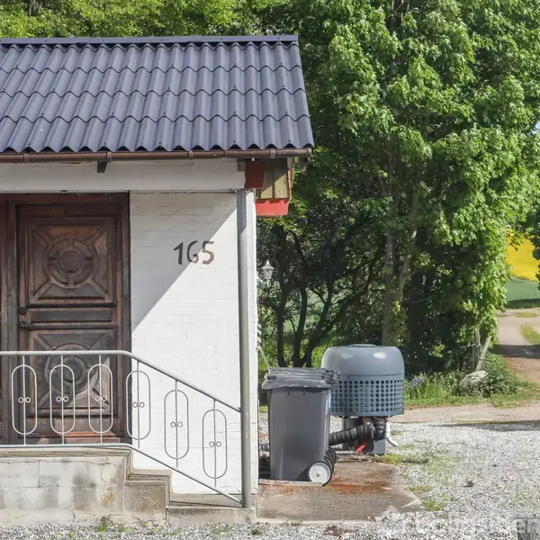Et hus med en brun trædør og nummeret 165 på facaden. En affaldsbeholder står foran, omgivet af træer. En grusvej løber langs huset, hvilket giver en landlig atmosfære.