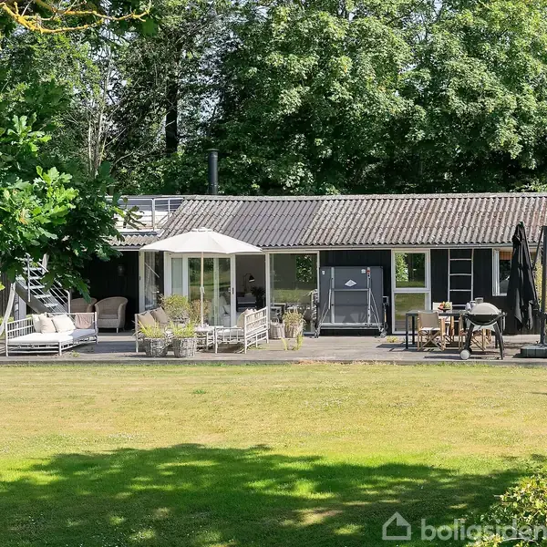 Et enkelt hus med terrasse ligger i en grøn have. På terrassen er der møbler, et lille bord og nogle planter. Stort træer omgiver området, hvilket skaber et naturskønt miljø.