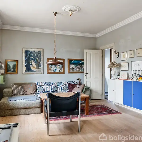 Stue med grå vægge. En sofa med puder står ved et vægophængt tv. Et sofabord foran sofaen, og en stol ved siden af. Væggen prydes af billeder og en skænk.