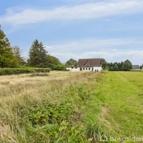 Et hvidt hus med rødt tag står isoleret. Omgivet af store græsarealer med marker og træer i baggrunden under en blå, skyet himmel.