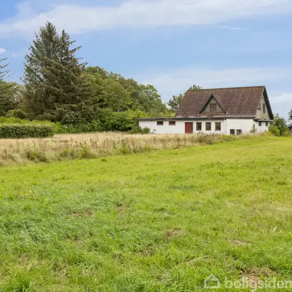 En hvid bygning med brunt tag står stille på en stor græsmark omgivet af træer under en blå himmel.