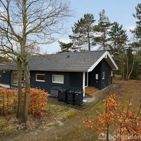 Et sort træhus står stille i en have med grussti omkranset af træer og buske. Et skilt foran huset viser teksten "TIL SALG".