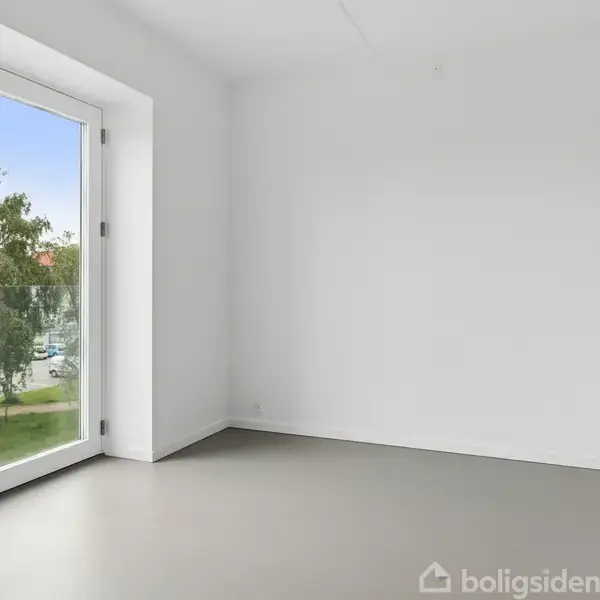Et tomt rum med hvide vægge og gulv, en stor glasdør til venstre åbner mod en udsigt med træer og en blå himmel udenfor.