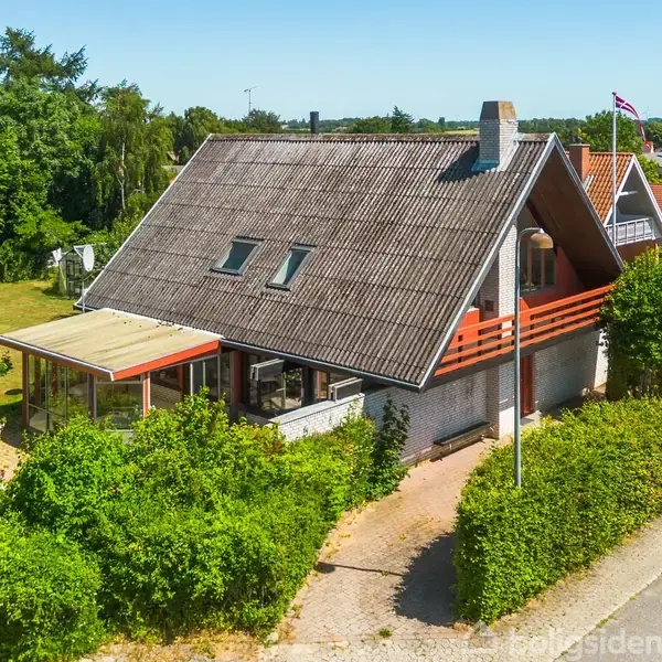 Et hus med asymmetrisk tag og to tagvinduer står omkranset af grønne buske. Det har en overbygget terrasse og ligger i et villakvarter med flere huse og træer omkring.