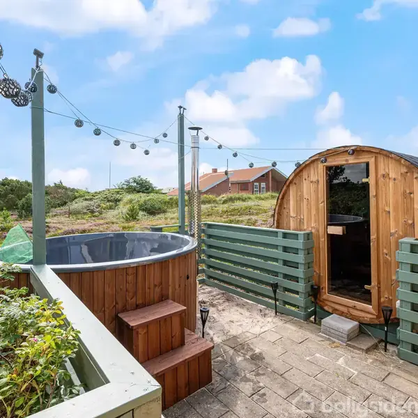 Udendørs spabad og sauna står i en terrassehave omgivet af hegn og planter. En lyskæde hænger over området, der ligger i et landskabeligt område med blåt himmelbillede.