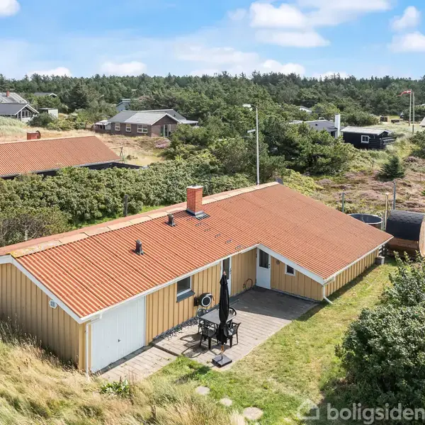 Et enfamiliehus med rødt tag står stille. Det er omgivet af grønne buske og andre huse ligger i baggrunden under en blå himmel med skyer.