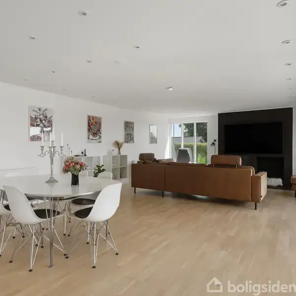 Spisebord med hvide stole står i en lys stue med lyse trægulve. Brun sofa vender mod en stor, mørk reol. Vægge har moderne kunst, og store vinduespartier giver udsigt.