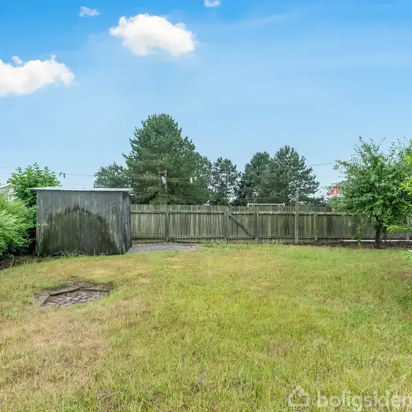 Et græsareal med et lille skur placeret til venstre. Omgivet af træer og en træhegn i baggrunden under en blå himmel med skyer.
