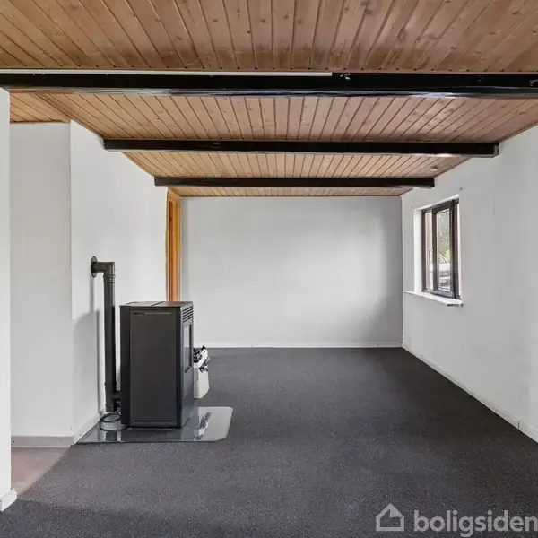 En brændeovn står stille på et mørkt tæppe i et rum med træloft og hvidmalede vægge; store vinduer til højre giver udsigt til omgivelserne udenfor.
