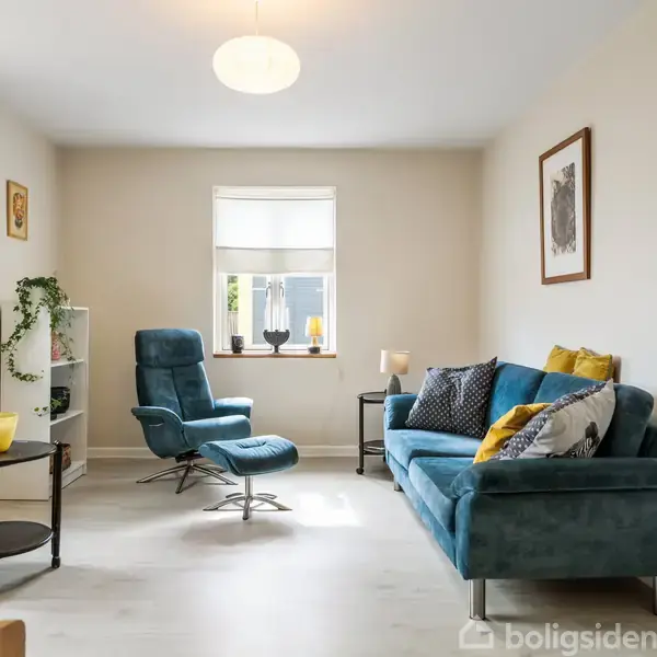 En stue med en blå sofa og matchende lænestol står mod en lys væg. Rummet har planter, puder, et lille rundt bord og en lampe.