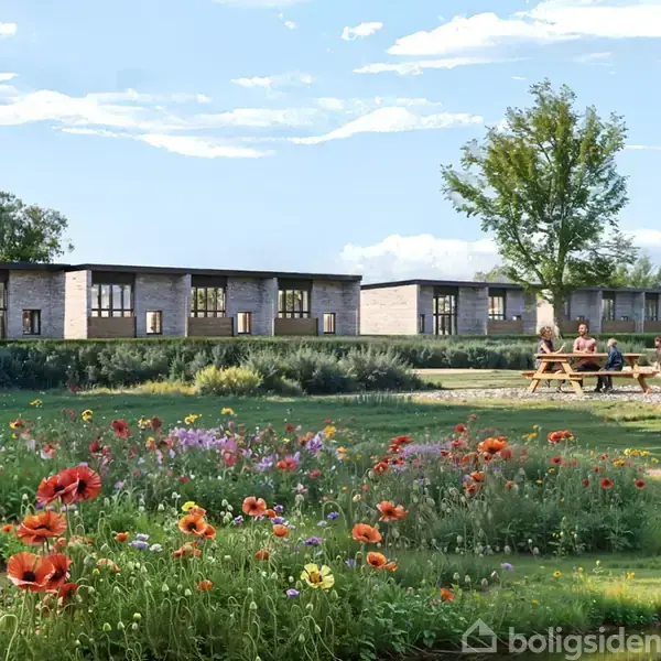 En familie sidder ved et picnicbord, omgivet af farverige blomster. I baggrunden ses en række moderne bygninger og et stort træ under en blå himmel med skyer.