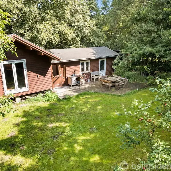 Et træhus står i en grøn have med græsplæne og skov. På terrassen foran huset er der et bord med stole. Sollys skinner gennem trætoppene.