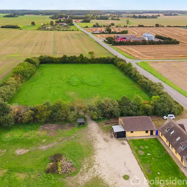 Gule bygninger med skrå tage ligger i en landlig gård, omgivet af grønne marker og træer, tæt på en vej. Fjernere ses åbne marker og spredte huse i horisonten.