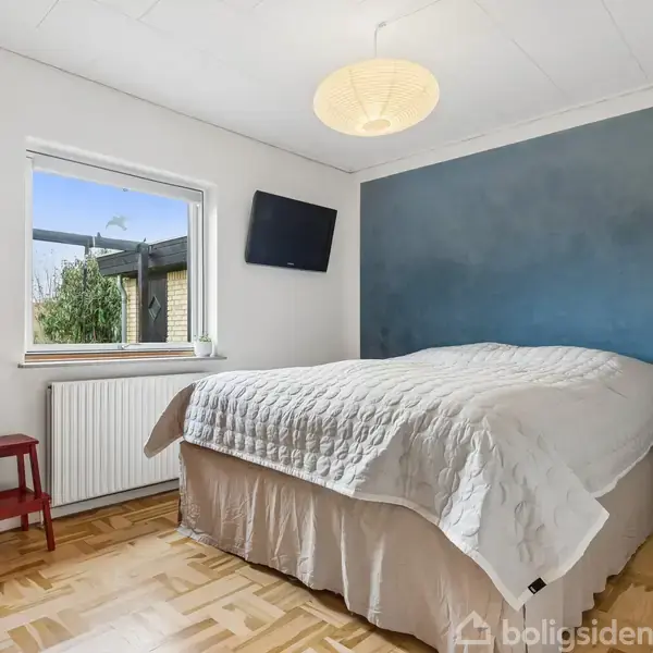 En seng med hvidt sengetæppe står i et lyst soveværelse med en blå accentvæg. En lampe hænger fra loftet, og et lille bord med en gul lampe står ved siden af sengen. En tv-skærm hænger på væggen ved siden af et vindue. En farverig plakat hænger på væggen.