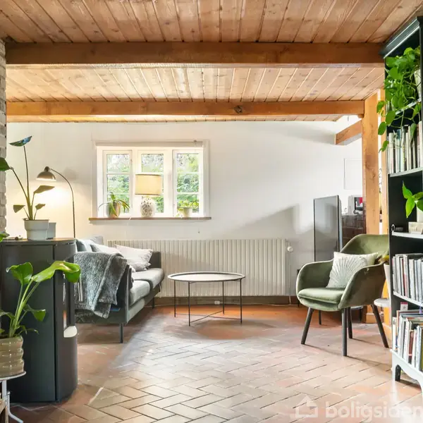 Stue med grønne planter, en sofa og stole omkring et sofabord; træloft og murstensvægge skaber en hyggelig atmosfære. En bogreol står til højre, vindue bagved.