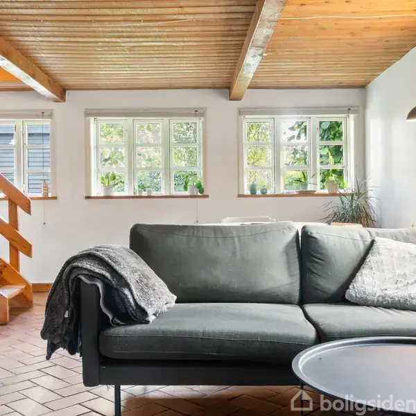 En grå sofa står i en lys stue med træloft og gulv. Ved siden af sofaen er der et rundt bord, en lampe og flere vinduer, der lader naturligt lys komme ind. I baggrunden ses en trappe.