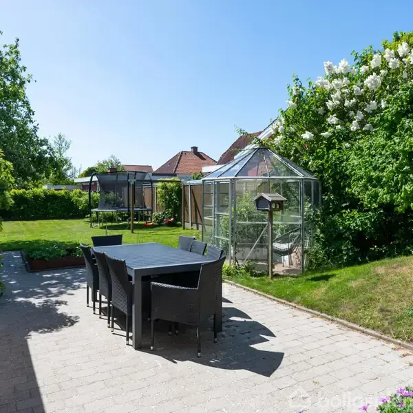 Et havebord med seks stole står på en flisebelagt terrasse. I baggrunden ses et drivhus omgivet af grønne buske og træer under en klar blå himmel.