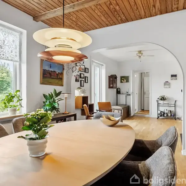 Et ovalt spisebord med en plante i midten står under en pendellampe. I baggrunden ses en lys stue med træloft og store vinduer med udsigt til en have.