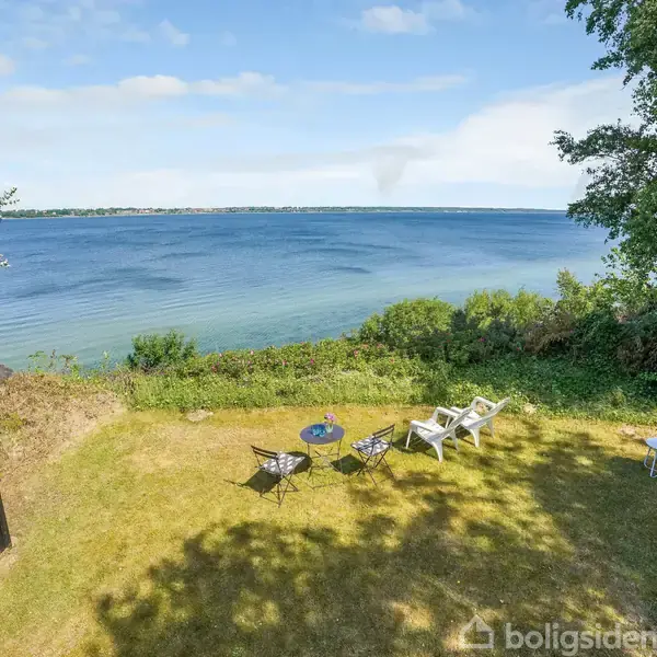 Et sommerhus står frit på en grund med udsigt til en blå sø eller hav. Lænestole og borde er placeret på græsplænen, hvilket skaber en afslappet atmosfære.