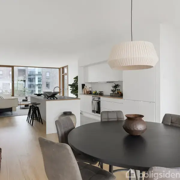 Spisestue med sort rundt bord omgivet af grå stole. Baggrunden viser et åbent køkken med hvide skabe. Store vinduer tilbyder udsigt til en moderne lejlighedsbygning. Ingen tekst fundet.