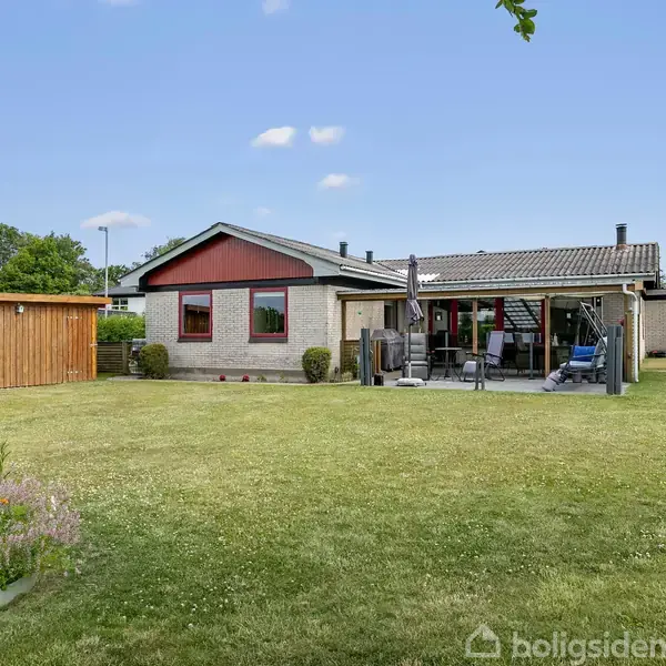 Hus med rødt tag og hvide vægge står i en rummelig grønt have. Et redskabsskur er til venstre, og en terrasse med møbler ses ved siden af huset.