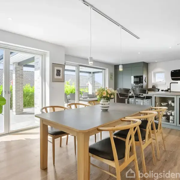 Et lyst køkken-alrum med et stort træbord og seks stole. Rummet har store vinduer med udsigt til en terrasse. Et køkkenområde med moderne apparater ses i baggrunden.