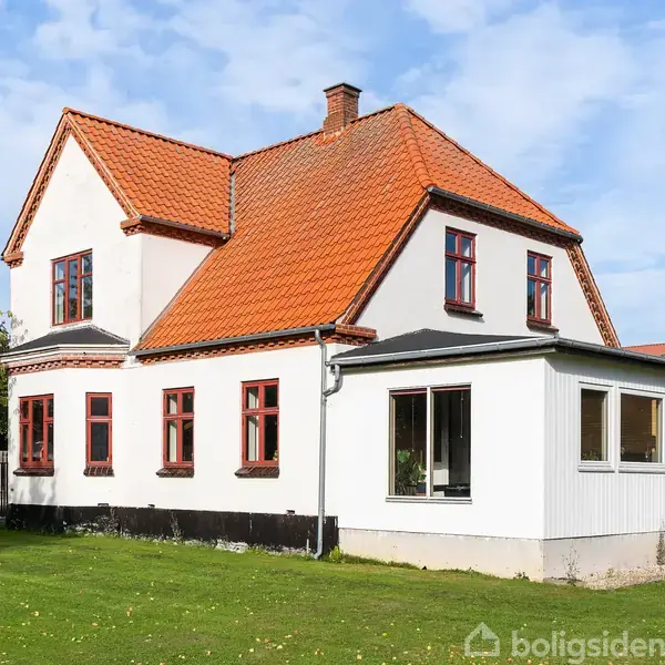 Et hvidt hus med rødt tegltag og røde vinduesrammer står på en grøn græsplæne. Huset har en nyere tilbygning og en blå himmel i baggrunden.
