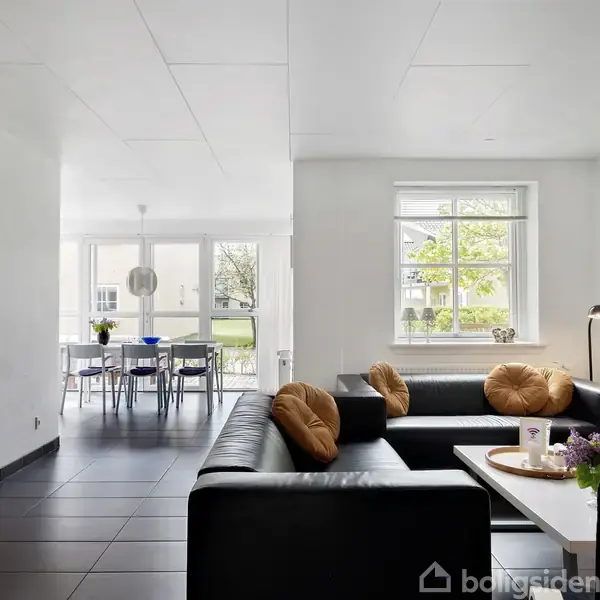 Sort sofa med brune puder står i en lys stue med et sofabord. Et spisebord med stole ses nær et vindue, og et maleri hænger på væggen.