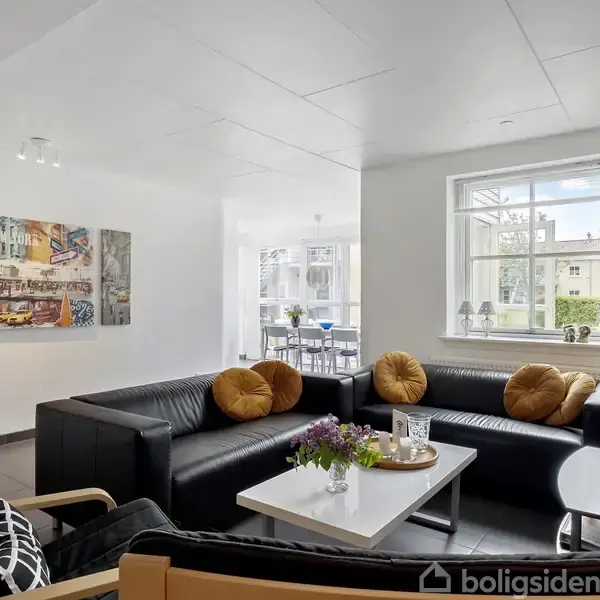 Sort sofaarrangement med gule puder står i en lys stue, udsmykket med maleri. Stort vindue skaber en lys atmosfære, med udsigt til en spisestue og have udenfor.