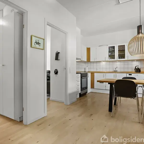 Spisebord med sorte stole står i et lyst køkken med hvide skabe og trædetaljer. En rund lampe hænger over bordet, og der er en åbning til en gang.