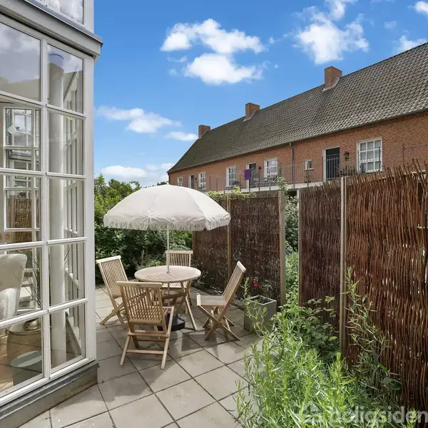 Terrassemøbler står på en flisebelagt gårdhave med et rundt bord og parasol, omringet af hegn og planter, beliggende ved siden af et hus med store vinduer.