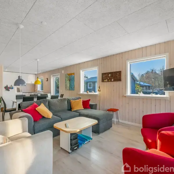 Stue med moderne sofagruppe og farverige puder dominerer rummet. TV hænger på væggen, mens vinduer slipper lys ind. Køkken ses i baggrunden. Møblerne er placeret på lyst trægulv.