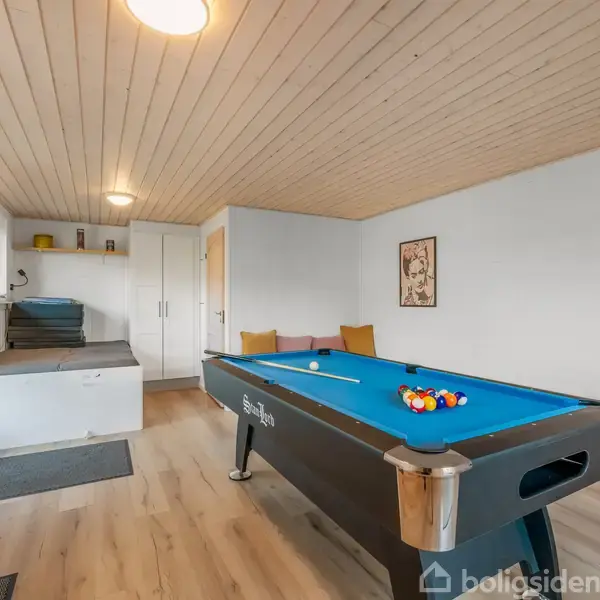 Et poolbord med blå filt står i et lyst rum med trægulv og træloft. Der er en dør til venstre, små hylder på væggen og et billede hænger til højre.