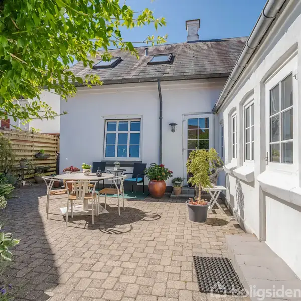 Hvidt hus med terrasse, indeholder bord og stole i midten. Store vinduer og planters sidder langs væggene. Træer og planter omgiver området, skaber en hyggelig gårdhave atmosfære.