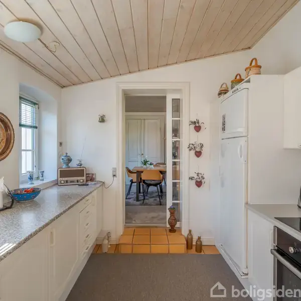 Køkken med hvide skabe og en marmor bordplade, hvorpå står køkkenredskaber. En dør fører til en spisestue med et bord og stole. Væggene har diverse pynt.