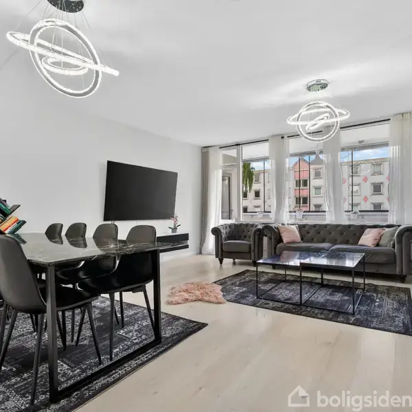 En moderne stue indeholder et langbord med sorte stole til venstre og to grå sofaer til højre. Store vinduer i baggrunden giver naturligt lys. Spiralformede loftlamper hænger over bordet og sofabordene. Mønstret tæppe ligger under bordene.